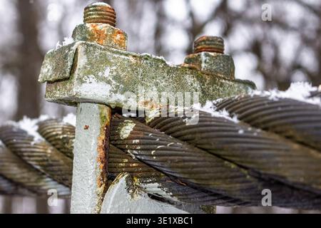 Weathered, snow-dusted suspension bridge cable with rusty industrial clamp and bolts. Close-up of durable metal structure in frosty winter landscape Stock Photo