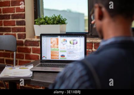 Back view of black male manager focused on business reports displayed on his laptop. African american businessman examining startup company presentation displayed on device screen at office desk. Stock Photo