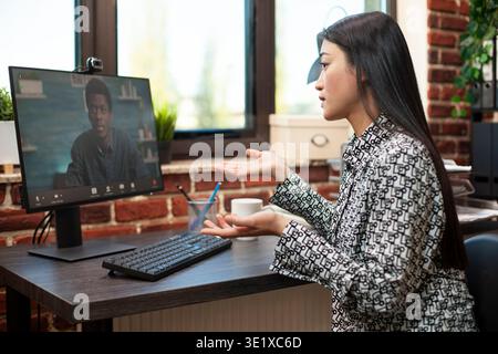Asian woman collaborating remotely on desktop pc, discussing project plans with colleague via virtual meeting. Female advisor engaging in video call to share strategies with african american client. Stock Photo