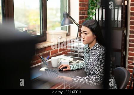 Female researcher near window types diligently, updating marketing analytics in brick wall office. Asian woman focuses on project updates at her desk, working on her computer in a modern workspace. Stock Photo