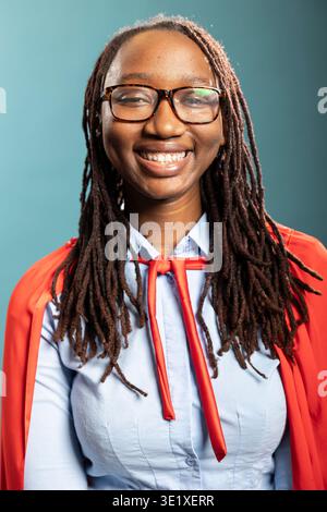 Closeup of young woman with cheerful expression poses confidently in red cape and blue shirt, symbolizing motivation and strength. Portrait of african american lady smiling brightly at camera. Stock Photo