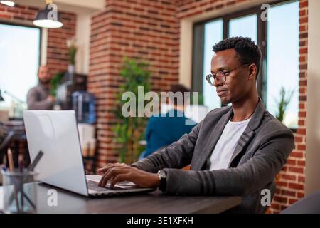 Male entrepreneur works diligently on financial research using his laptop in startup office. Black businessman at desk with his personal computer is focused on improving company status growth. Stock Photo