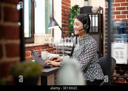 Female employee uses desktop pc, typing on keyboard and engaging in a lively conversation through wired headphones in modern workplace. Smiling woman seated at office desk, using computer and headset. Stock Photo