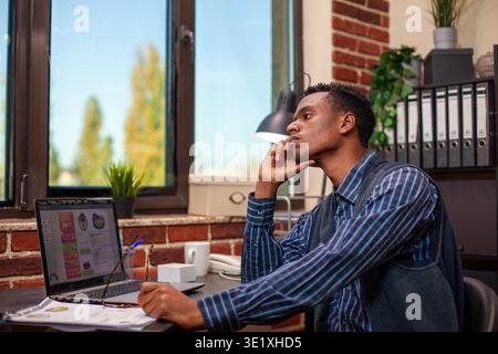Tired male analyst holding his chin at desk and staring outside the window in brick wall workspace. Manager leans head on his hand, feeling exhausted by business analytics research at startup office. Stock Photo