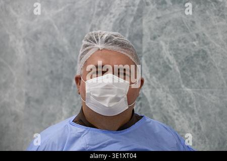 Portrait of professional ophthalmologist Dr. Ergun Rashidalizade wearing a medical mask, cap, and blue scrubs against a marble background in a clinic. Stock Photo
