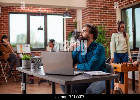 White business manager sitting at table with laptop, deep in thought, holding a pen to his chin. Male entrepreneur with a beard, reflecting on company projects as he looks around brick wall workplace. Stock Photo