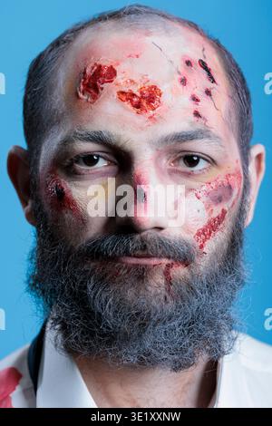 Portrait of actor dressed as horror movie zombie with fake blood and scars on face. Close up of man costumed as undead monster covered in bruises, professional makeup, isolated over studio background Stock Photo