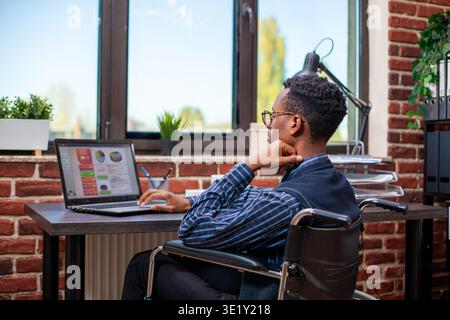 Black businessman in wheelchair studying digital financial documents on laptop. African american male analyst recovering from physical trauma and reviewing company strategy ideas on device in office. Stock Photo