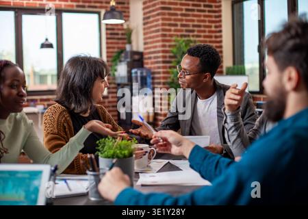 Businessman leads a group discussion with employees in startup office. Caucasian woman and black male analyst exchange marketing strategy ideas during company team meeting in brick wall workspace. Stock Photo