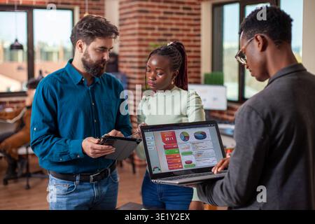 Businessman and woman reviewing online documents on tablet as black man checking business analytics on his laptop. Male and female coworkers using digital devices in brick wall office. Stock Photo