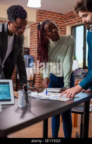African american female manager with pen in hand, speaking to male employees in brick wall office. Businesswoman points at documents, having company project discussion with colleagues at table. Stock Photo