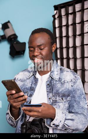 Black male photographer holding a credit card and smartphone, completing an online transaction for his photography equipment. African American cameraman uses his mobile device to verify the payment. Stock Photo