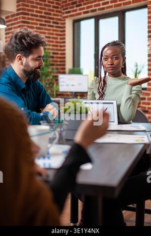 Black entrepreneur using tablet to outline company plans to colleagues during discussion in modern workspace. Female financial advisor speaking to startup team and showing charts on device in meeting. Stock Photo