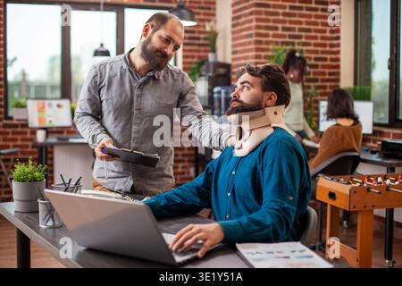 Injured analyst seated at desk with his laptop, receiving friendly support from coworker in office. Male manager stands with clipboard and speaks to his employee with a neck brace in modern workspace. Stock Photo