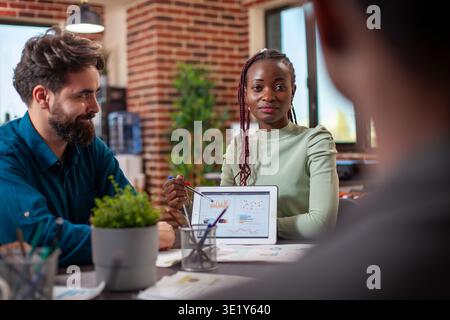 Black entrepreneur guiding her colleagues with market strategy insights, utilizing a digital device. Female startup leader using a tablet to explain company goals to her team in brick wall office. Stock Photo