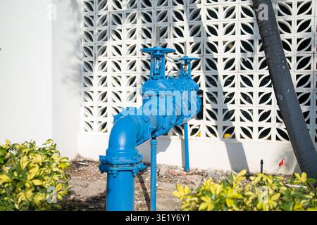 Heavy-duty blue-painted iron water valves and piping system located outside a building with a white-patterned block wall behind. Stock Photo