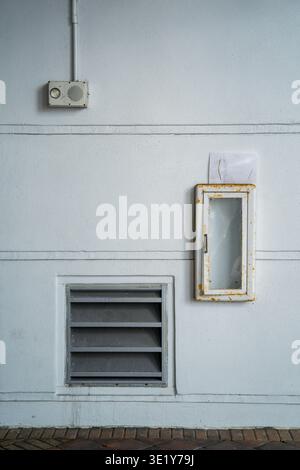 Front view of a white textured exterior building wall with a ventilation grill and an old rusty fire alarm system component. Stock Photo
