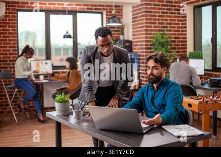 Caucasian man with laptop reviews online documents with african american coworker at table in modern workspace. Male colleagues remain focused on achieving business objectives using digital device. Stock Photo