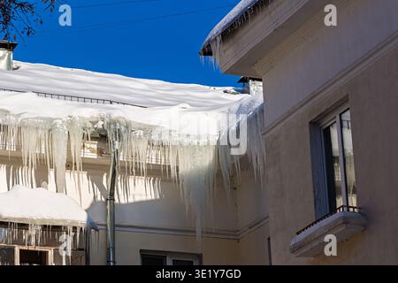 Dramatic, huge icicles gleam on a thick blanket of rooftop snow against a vibrant winter sky, showcasing nature's cold beauty and potential danger Stock Photo