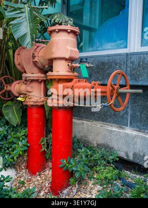 Red fire department connection pipeline with multiple manual gate valves installed in a garden bed near a modern building with blue glass windows. Stock Photo