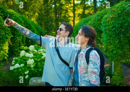 Two young men are taking a selfie with a smartphone, standing in a lush green garden tunnel with white hydrangea flowers in the park on a sunny day. Stock Photo