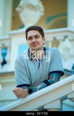 Portrait of a young man with tattoos looking up and leaning on a white railing of a classical building in the summer garden during a bright day. Stock Photo