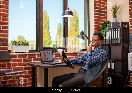 Black male advisor with clipboard reading project update to client on phone call. African american manager sitting in brick wall office, using landline to speak with his employees about business plan. Stock Photo