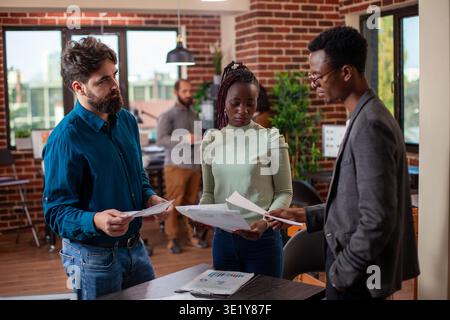 Startup employees stand around a table, reviewing company growth ideas and analyzing data in collaborative setting. Male and female colleagues holding documents and brainstorming strategies in office. Stock Photo