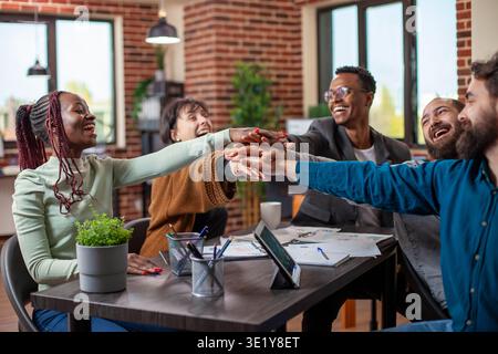 Multiethnic colleagues cheerfully celebrating completion of business project. Diverse startup team seated around table, stacking hands together with excitement, teamwork concept in modern office. Stock Photo