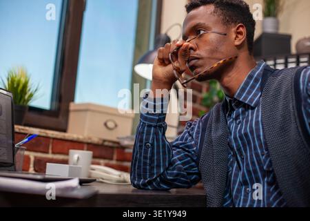 Overwhelmed male analyst holding his eyeglasses and looking outside the window at startup company workspace. Businessman with spectacles in hand, sitting at desk, deeply thinking about strategy ideas. Stock Photo