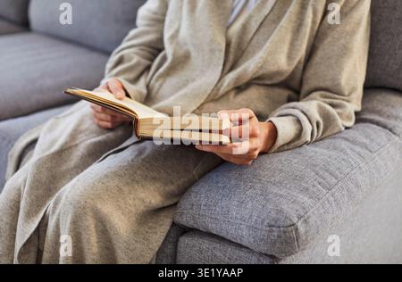 Faceless person sitting on sofa at home reading a book in living room enjoying literature indoors. Stock Photo