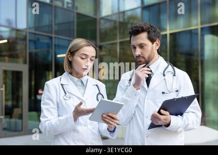 Two medical professionals, a man and a woman wearing lab coats and stethoscopes, sharing and analyzing information on a digital tablet in front of a modern hospital building Stock Photo