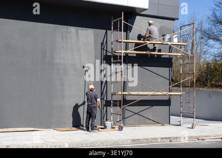 Workers painting an exterior building wall using rollers and scaffolding on a bright day. Copy space Stock Photo
