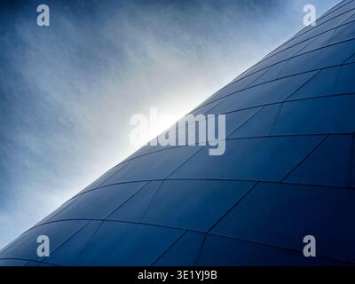 A glimpse of sun flares through a very light cloud cover as it rises over the horizon formed by a exterior skin of a modern building. Stock Photo