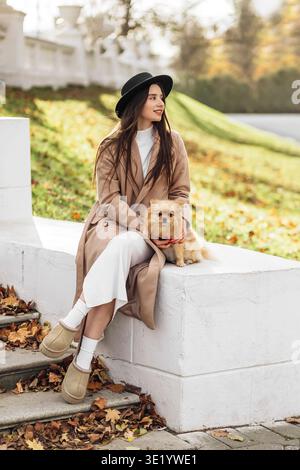 Woman with chestnut hair in black hat and beige coat hugging her beloved Pomeranian dog on a blurred autumn-spring background, sunlit, animal love, pe Stock Photo