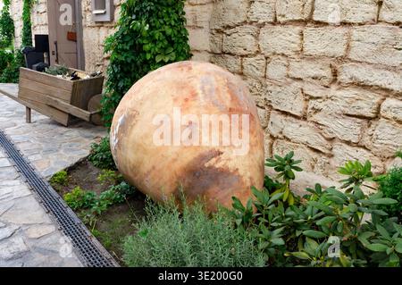 A large, brown, ceramic vase is sitting on the ground next to some plants. The vase is in the middle of a garden and is surrounded by green plants Stock Photo