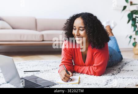 Young Woman Studies at Home While Taking Notes on a Notepad in Front of a Laptop Stock Photo