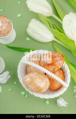 Creative spring holiday composition. Easter eggs dyed with natural onion skins in a white basket and stands, white tulips and bunny figurines on a gre Stock Photo