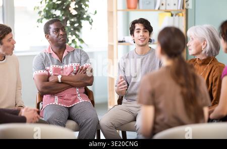 Male and female colleagues of various ages and nationalities talking sitting in circle on chairs in office lounge during break Stock Photo