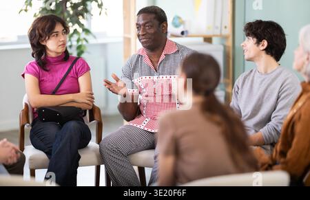 Male and female colleagues of various ages and nationalities talking sitting in circle on chairs in office lounge during break Stock Photo