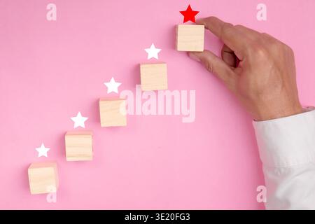 Hand placing wooden cube with red star on top of stack representing achievement goal and success on pink background Stock Photo