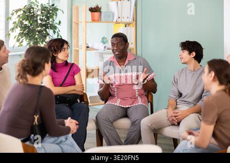 Male and female colleagues of various ages and nationalities talking sitting in circle on chairs in office lounge during break Stock Photo