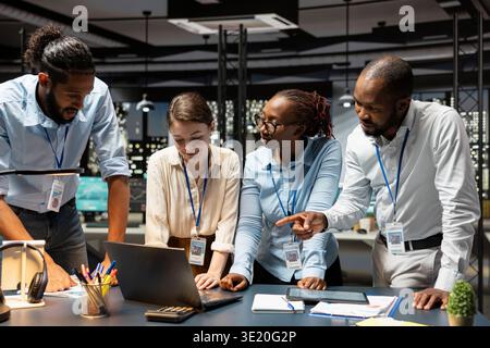 Multiethnic team of office clerks collaborating on completing spreadsheets using devices, analyzing data. Diverse coworkers researching information online on notebook and tablet, working overnight Stock Photo