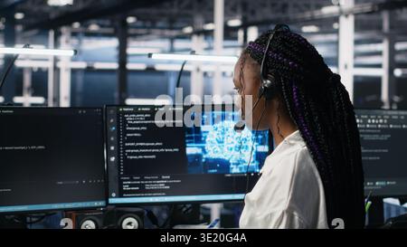 Admin in data center wearing headset assisting users with artificial intelligence troubleshooting. African american woman in server room hub managing support tickets using automated systems, camera A Stock Photo