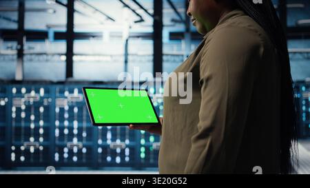 Close up of technician at work using green screen tablet in data center to check hardware for system stability. Employee using mockup device to oversee server farm and configure equipment, camera A Stock Photo