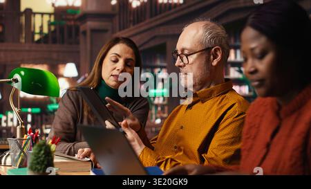 Mature college students completing class notes on papers and tablet, writing important information in a study area under green lamp. Colleagues pursuing academic qualifications. Stock Photo