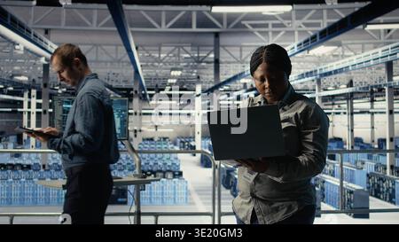 Data center technician deploys machine learning software on infrastructure racks. African american woman in server farm trains AI systems with datasets using laptop to support applications, camera A Stock Photo