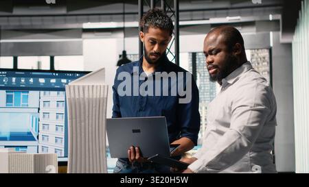 Multiethnic group of developers and contractors discussing urban housing models and blueprints layout revisions for upcoming residential development project. Smart city planning session. Stock Photo