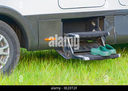 A pair of green rubber clogs placed on the retractable step of a recreational vehicle parked on green grass, highlighting outdoor travel and camping l Stock Photo