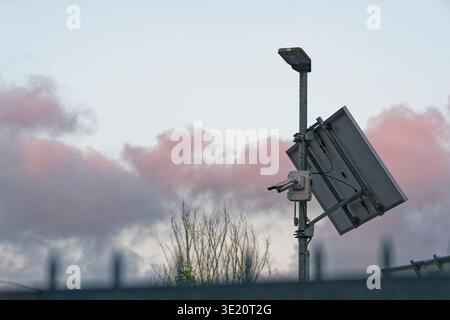 A solar panel mounted on a pole powers a street light and a security camera with a backdrop of pink and gray clouds during dusk. Stock Photo